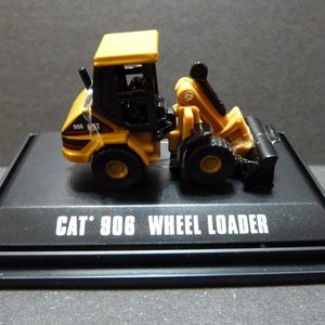 May include: A yellow and black CAT 906 Wheel Loader model on a black display stand. The model features a detailed design with a front bucket and a cab. The text "CAT 906 WHEEL LOADER" is printed on the stand.
