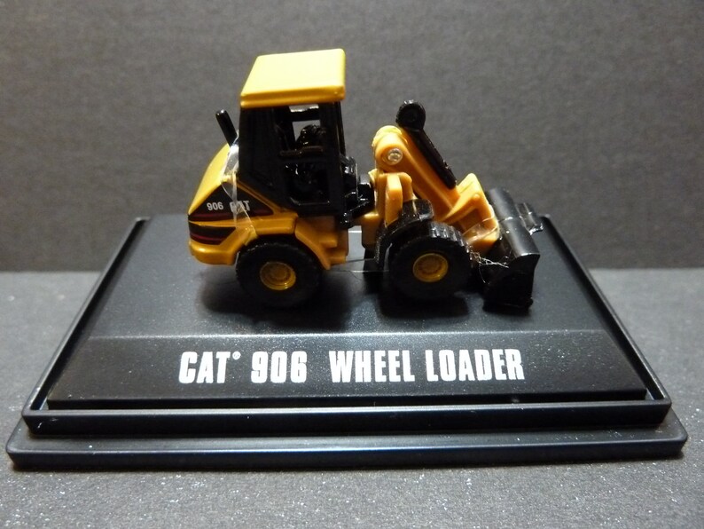 May include: A yellow and black CAT 906 Wheel Loader model on a black display stand. The model features a detailed design with a front bucket and a cab. The text "CAT 906 WHEEL LOADER" is printed on the stand.