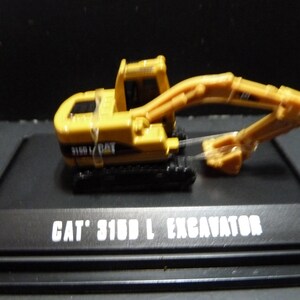 May include: A yellow CAT 315D L excavator model on a black display stand. The model features a detailed design with a movable arm and bucket. The text "CAT 315D L EXCAVATOR" is printed in white on the stand.