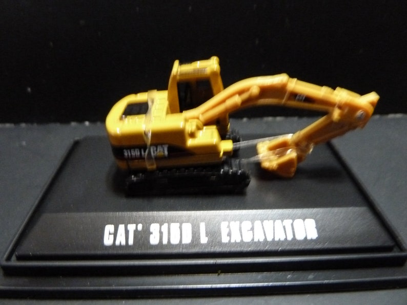 May include: A yellow CAT 315D L excavator model on a black display stand. The model features a detailed design with a movable arm and bucket. The text "CAT 315D L EXCAVATOR" is printed in white on the stand.