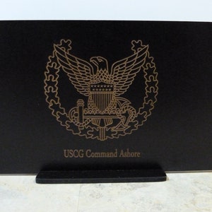 May include: A black wooden plaque with a gold-colored US Coast Guard emblem and the text "USCG Command Ashore".