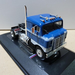 May include: A blue cab-over semi-truck model featuring a chrome grille, vertical exhaust stack, and roof-mounted lights. The truck has a white stripe along the side, silver fuel tanks, and black wheels, all mounted on a black display base.