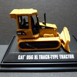 May include: A yellow Caterpillar D5G XL track-type tractor model. The miniature bulldozer features a blade, tracks, and a cab. The model is displayed on a black base with the text "CAT D5G XL TRACK-TYPE TRACTOR".