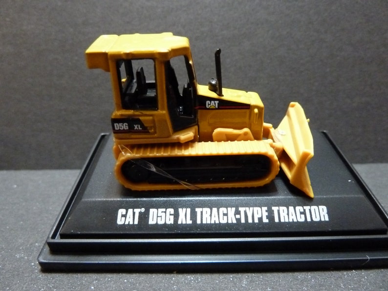 May include: A yellow Caterpillar D5G XL track-type tractor model. The miniature bulldozer features a blade, tracks, and a cab. The model is displayed on a black base with the text "CAT D5G XL TRACK-TYPE TRACTOR".