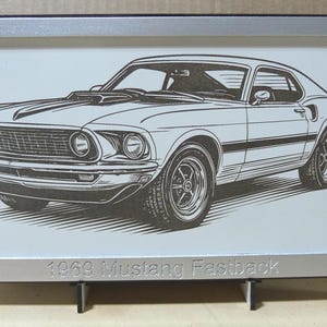 May include: Framed black and white illustration of a 1969 Mustang Fastback. The artwork is in a silver frame and includes the text "1969 Mustang Fastback" at the bottom. The car is depicted in a detailed, vintage style.