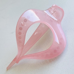 May include: A translucent light pink hair clip with a curved, open-frame design. The accessory features a smooth, glossy finish and a series of small, circular indentations along the outer edges of the two main arched prongs that meet at a point.
