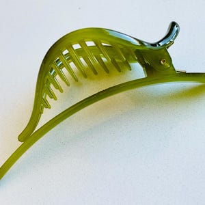 May include: A translucent olive green hair clip featuring a curved, arched design with a series of parallel teeth along the top edge. The accessory includes a hinged mechanism with a small metal pin, allowing the two main sections to open and close.