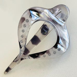 May include: A translucent hair clip with a teardrop-shaped frame featuring a marbled pattern of grey, black, and clear tones. The accessory has a smooth, glossy finish and a hinged mechanism with a series of small circular indentations along the edges.