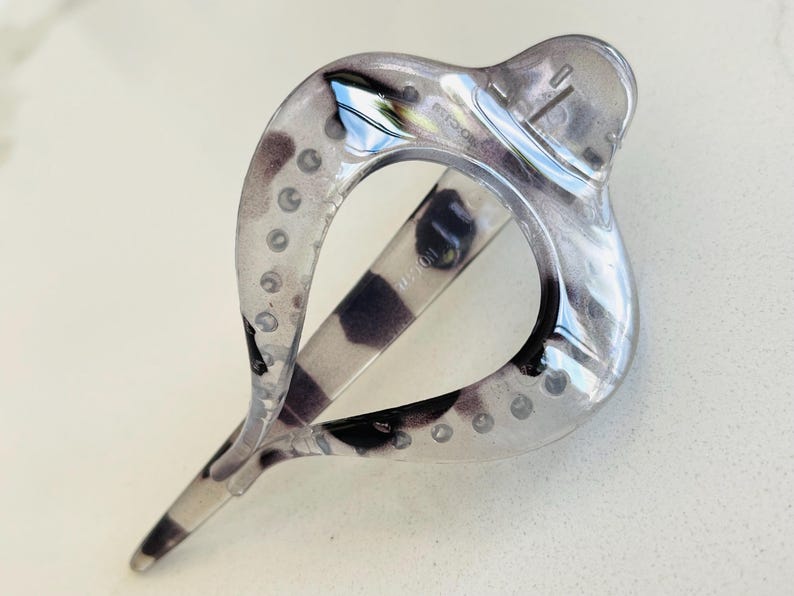 May include: A translucent hair clip with a teardrop-shaped frame featuring a marbled pattern of grey, black, and clear tones. The accessory has a smooth, glossy finish and a hinged mechanism with a series of small circular indentations along the edges.