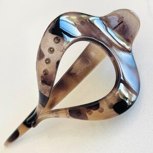 May include: A translucent brown and black tortoiseshell hair clip with a teardrop-shaped open center. The accessory features a smooth, glossy finish and a curved, elongated design with a pointed end, showcasing a mottled pattern throughout the material.
