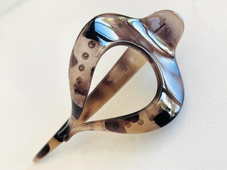 May include: A translucent brown and black tortoiseshell hair clip with a teardrop-shaped open center. The accessory features a smooth, glossy finish and a curved, elongated design with a pointed end, showcasing a mottled pattern throughout the material.