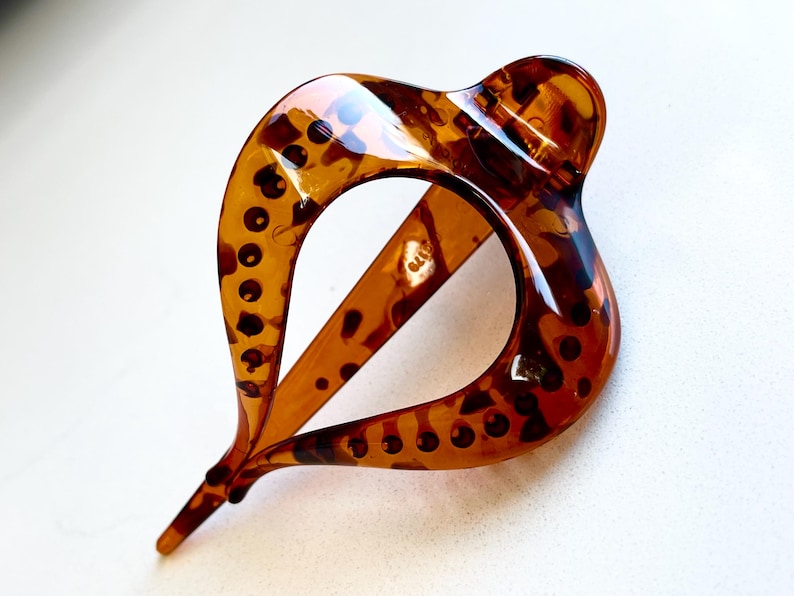 May include: A teardrop-shaped hair clip featuring a glossy, translucent amber and dark brown tortoiseshell pattern. The accessory has a smooth, curved frame with a central open space and a pointed tip, showcasing a polished finish throughout.