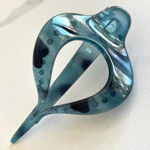 May include: A translucent teal hair clip with a teardrop-shaped frame and a mottled dark blue pattern. The accessory features a hinged mechanism at the top and a pointed tail at the bottom, constructed from a smooth, glossy plastic material.