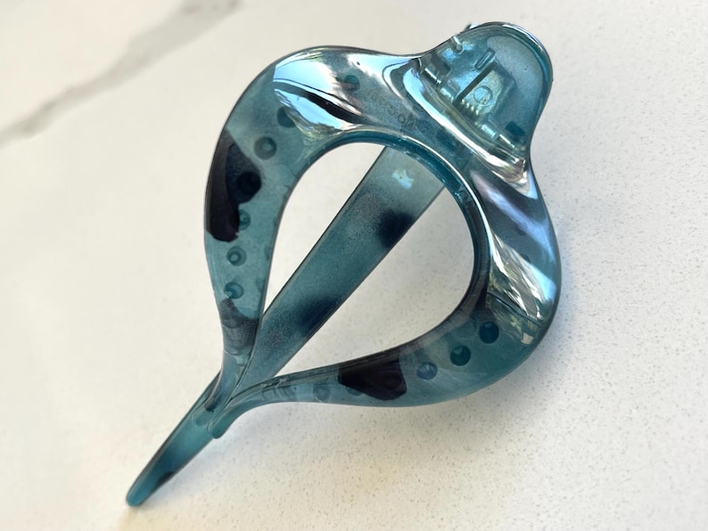 May include: A translucent teal hair clip with a teardrop-shaped frame and a mottled dark blue pattern. The accessory features a hinged mechanism at the top and a pointed tail at the bottom, constructed from a smooth, glossy plastic material.