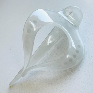May include: A translucent white hair claw clip with a curved, teardrop-shaped frame. The clip features a series of small, circular cutouts along the outer edges and a spring-loaded hinge mechanism at the top for securing hair in place.