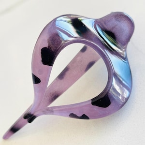 May include: A translucent purple hair clip with a teardrop-shaped opening and irregular black spots throughout the material. The accessory features a smooth, glossy finish and a pointed end, designed with a curved, open frame structure.