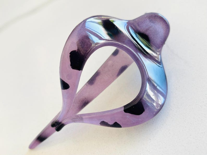 May include: A translucent purple hair clip with a teardrop-shaped opening and irregular black spots throughout the material. The accessory features a smooth, glossy finish and a pointed end, designed with a curved, open frame structure.