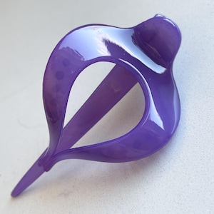 May include: A glossy, translucent purple hair clip featuring a curved, teardrop-shaped frame with a pointed end and a rounded top section. The accessory has a smooth, reflective surface and a hollow center, designed for securing hair in place.