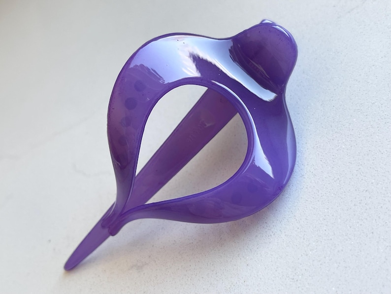 May include: A glossy, translucent purple hair clip featuring a curved, teardrop-shaped frame with a pointed end and a rounded top section. The accessory has a smooth, reflective surface and a hollow center, designed for securing hair in place.