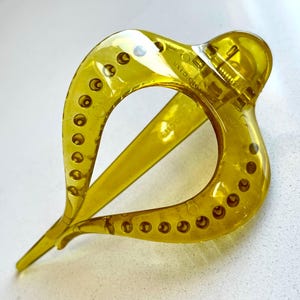 May include: A translucent olive green hair claw clip with a teardrop-shaped frame. The clip features a series of small circular indentations along the outer edges of the frame and a spring-loaded hinge mechanism at the top for securing hair.