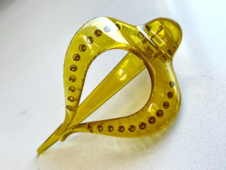 May include: A translucent olive green hair claw clip with a teardrop-shaped frame. The clip features a series of small circular indentations along the outer edges of the frame and a spring-loaded hinge mechanism at the top for securing hair.