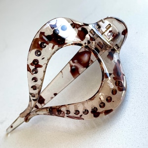 May include: A translucent hair claw clip featuring a brown and black tortoiseshell pattern. The clip has a curved, open-loop design with a spring-loaded hinge mechanism at the top and a pointed tip at the bottom, showcasing a glossy finish.