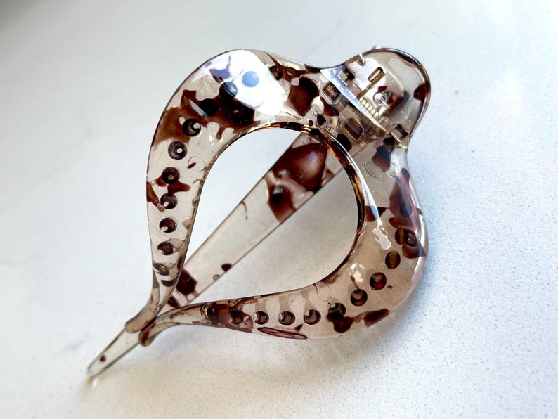 May include: A translucent hair claw clip featuring a brown and black tortoiseshell pattern. The clip has a curved, open-loop design with a spring-loaded hinge mechanism at the top and a pointed tip at the bottom, showcasing a glossy finish.