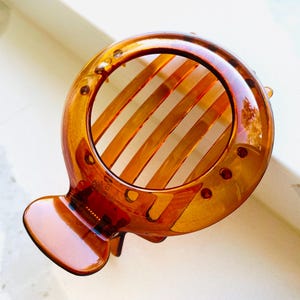 May include: A tortoiseshell-colored hair claw clip. The clip has a circular design with parallel bars and a rounded base. The clip is made of a glossy, translucent material and has a smooth finish. The clip is designed to hold hair in place.