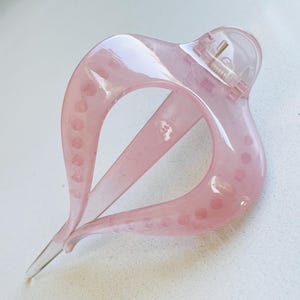May include: A translucent light pink hair claw clip with a curved, teardrop-shaped frame. The accessory features a spring-loaded hinge mechanism at the top and a series of small, circular indentations along the inner edges of the two main prongs.