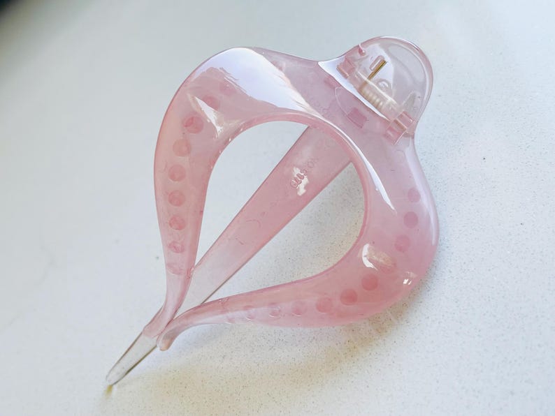 May include: A translucent light pink hair claw clip with a curved, teardrop-shaped frame. The accessory features a spring-loaded hinge mechanism at the top and a series of small, circular indentations along the inner edges of the two main prongs.