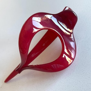 May include: A glossy, deep red hair clip with a smooth, curved, open-frame design. The accessory features a pointed end and a rounded, fluid shape that creates a hollow center, showcasing a polished, reflective surface throughout the entire piece.