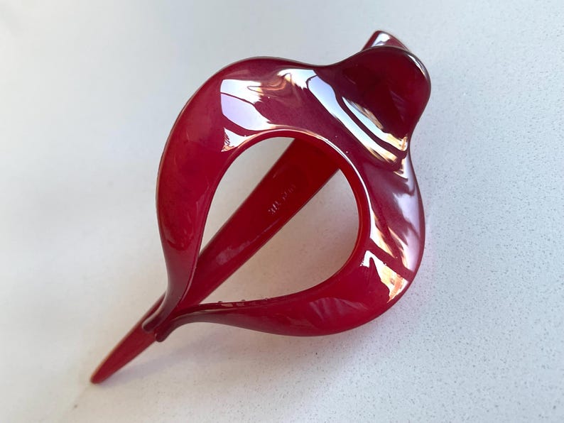May include: A glossy, deep red hair clip with a smooth, curved, open-frame design. The accessory features a pointed end and a rounded, fluid shape that creates a hollow center, showcasing a polished, reflective surface throughout the entire piece.