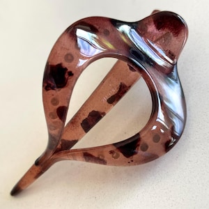 May include: A translucent, dark brown hair clip with a glossy finish and a teardrop-shaped cutout. The material features a mottled, tortoiseshell-like pattern with darker brown spots and lighter, semi-transparent areas throughout the structure.