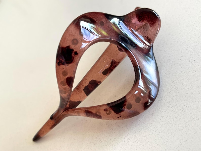 May include: A translucent, dark brown hair clip with a glossy finish and a teardrop-shaped cutout. The material features a mottled, tortoiseshell-like pattern with darker brown spots and lighter, semi-transparent areas throughout the structure.