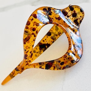 May include: A hair clip with a teardrop-shaped frame featuring a translucent amber base and a speckled pattern of dark brown and black spots. The accessory has a smooth, glossy finish and a pointed end, showcasing a distinct open center design.