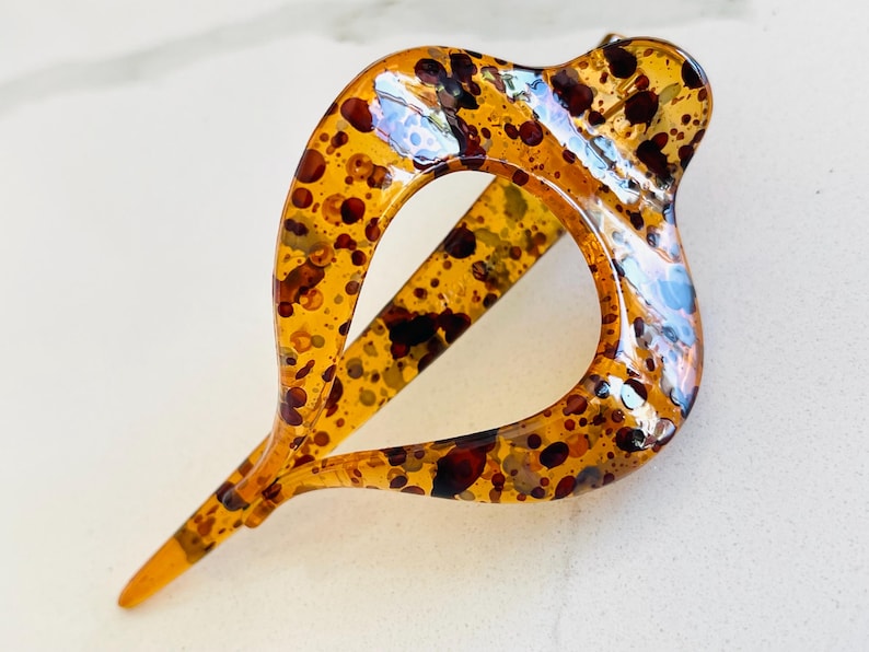 May include: A hair clip with a teardrop-shaped frame featuring a translucent amber base and a speckled pattern of dark brown and black spots. The accessory has a smooth, glossy finish and a pointed end, showcasing a distinct open center design.