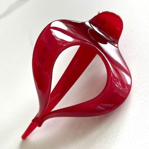 May include: A glossy, red hair clip with a unique, abstract design. The clip features a curved, flowing shape with a central opening and a pointed end. The hair accessory is made of a shiny material.
