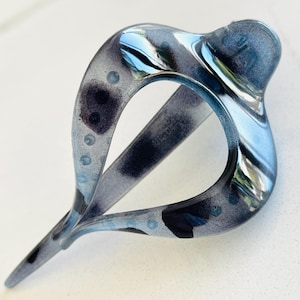 May include: A teardrop-shaped hair clip featuring a glossy, translucent finish with a marbled pattern of dark gray, black, and light blue tones. The accessory has a smooth, curved frame with a pointed end and a central open space for styling.