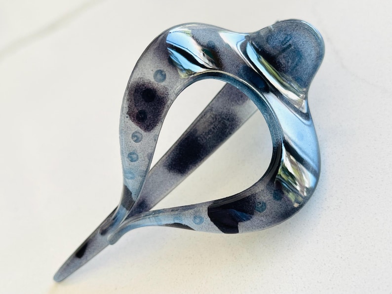 May include: A teardrop-shaped hair clip featuring a glossy, translucent finish with a marbled pattern of dark gray, black, and light blue tones. The accessory has a smooth, curved frame with a pointed end and a central open space for styling.