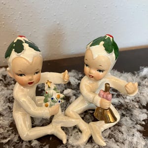 May include: Two vintage ceramic Christmas figurines. Each figurine is a cherubic figure with a green and white holly leaf hat and a red berry. One holds a small Christmas tree, the other a gold bell with a pink handle.