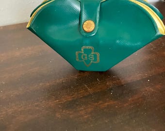 Könnte beinhalten: Ein dunkelgrünes, halbstarres Brillenetui in Muschelform mit einem goldfarbenen Druckknopfverschluss. Auf der Vorderseite befindet sich ein goldenes, geprägtes Logo mit den Buchstaben GS in einer Kleeblattform. Das Innenfutter ist in Gelb gehalten.