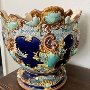 May include: A ceramic planter featuring a scalloped rim and a complex relief pattern. The surface displays a combination of deep cobalt blue, light turquoise, and warm brown glazes, with a textured geometric background and ornate scrollwork.