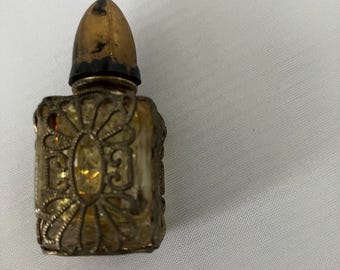 May include: An antique perfume bottle with a gold-colored, ornate design. The bottle has a rectangular shape with a decorative floral pattern and a gold-colored, bullet-shaped stopper. The bottle is likely made of glass and metal.