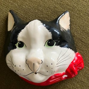 May include: A black and white ceramic cat head wall decor with a red bow. The cat has green eyes and a white nose.