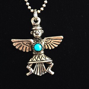 May include: Silver-toned pendant necklace featuring a stylized figure with outstretched wings and a turquoise-colored center stone. The pendant hangs from a ball chain, creating a striking contrast against the dark background.