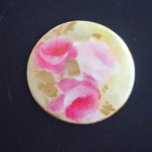 May include: A round, cream-colored ceramic button with a painted design of pink roses on a yellow background.