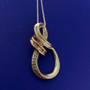May include: Silver-toned pendant necklace with an infinity symbol design. The pendant features a combination of silver and gold-toned metal, with small, sparkling accents. The necklace hangs on a delicate silver chain against a blue background.