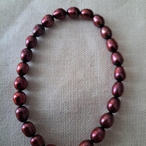 May include: A circular bracelet composed of numerous dark maroon, oval-shaped pearls strung together. The pearls exhibit a smooth, lustrous surface with subtle variations in their deep reddish-brown color, forming a continuous loop of beads.