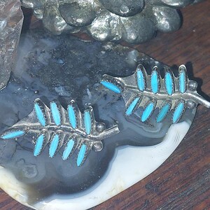 May include: A pair of silver-toned leaf-shaped earrings with turquoise-colored stone accents. The earrings are displayed on a gray and white stone surface, with other stones in the background. The earrings have a detailed, textured design.