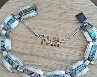 May include: A silver bracelet with a chain clasp and rectangular abalone shell inlays. The bracelet is on a wooden surface.